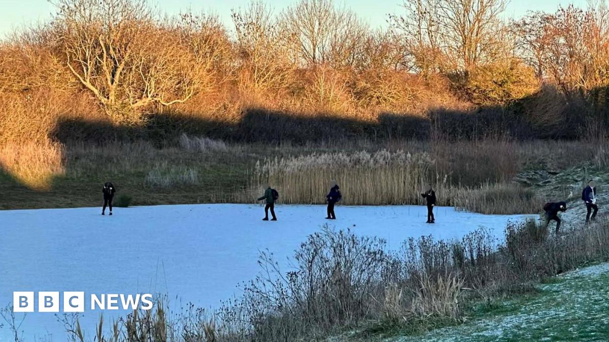 Four children attempt to cross the frozen Sherrer Pond in Swindon with others pictured nearby. The pond is surrounded by reeds, plants and trees and the grass around it is covered with ice.