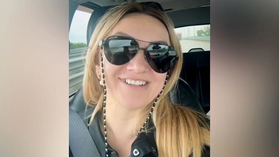 Polina Azarnykh looking straight at the camera while seated in a vehicle. She has long blonde hair and is wearing large sunglasses and a black top and is smiling.
