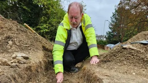 Bedford Roman Villa Project Steve Cocking, kneeling down with mud all around him. There are tools to his left. He is holding something. He is wearing a high-vis jacket, black trousers, white shirt and black top. He has a moustache and short fair hair. A building is behind him and trees are all around him. 