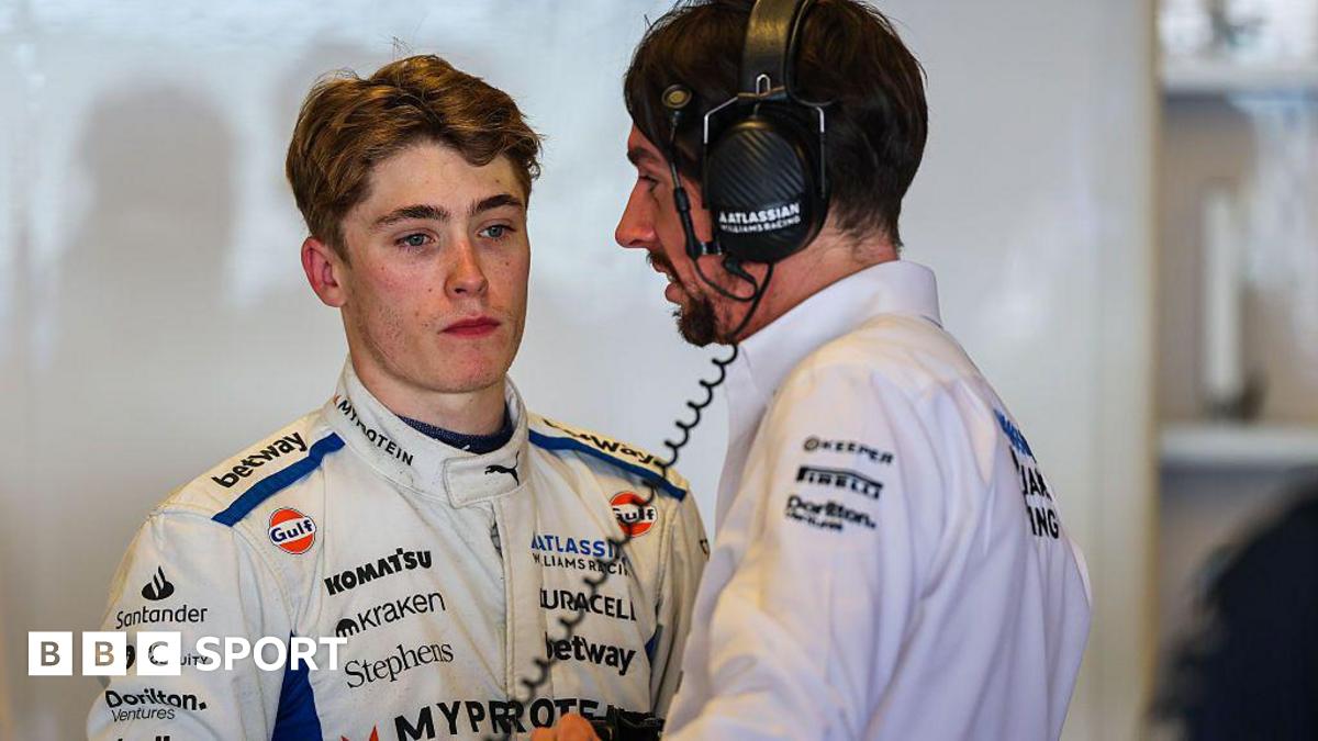 Luke Browning in the garage during F1 testing with Williams Racing, wearing a largely white race suit featuring several sponsor logos