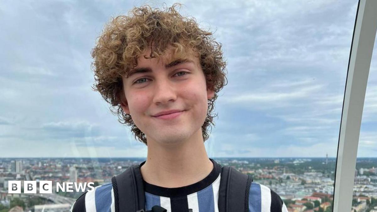Ethan Brown looking directly at the camera. He has curly brown hair and is wearing a t-shirt with blue, black and white vertical stripes, and has a rucksack on his back. In the background is a view over a city.