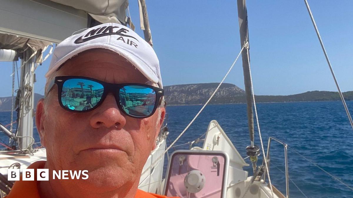 Middle-aged man on a sailing boat faces the camera wearing black sunglasses with reflective lenses, an orange top and white cap