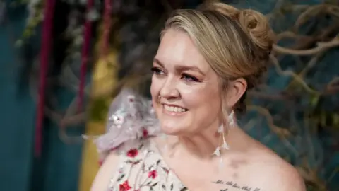 Getty Images Colleen Hoover at a film premiere with her blonde hair in an up-do, wearing dangly white feather earrings - she has a one-shoulder jewelled white dress on, and a tattoo of writing can be seen on her shoulder 