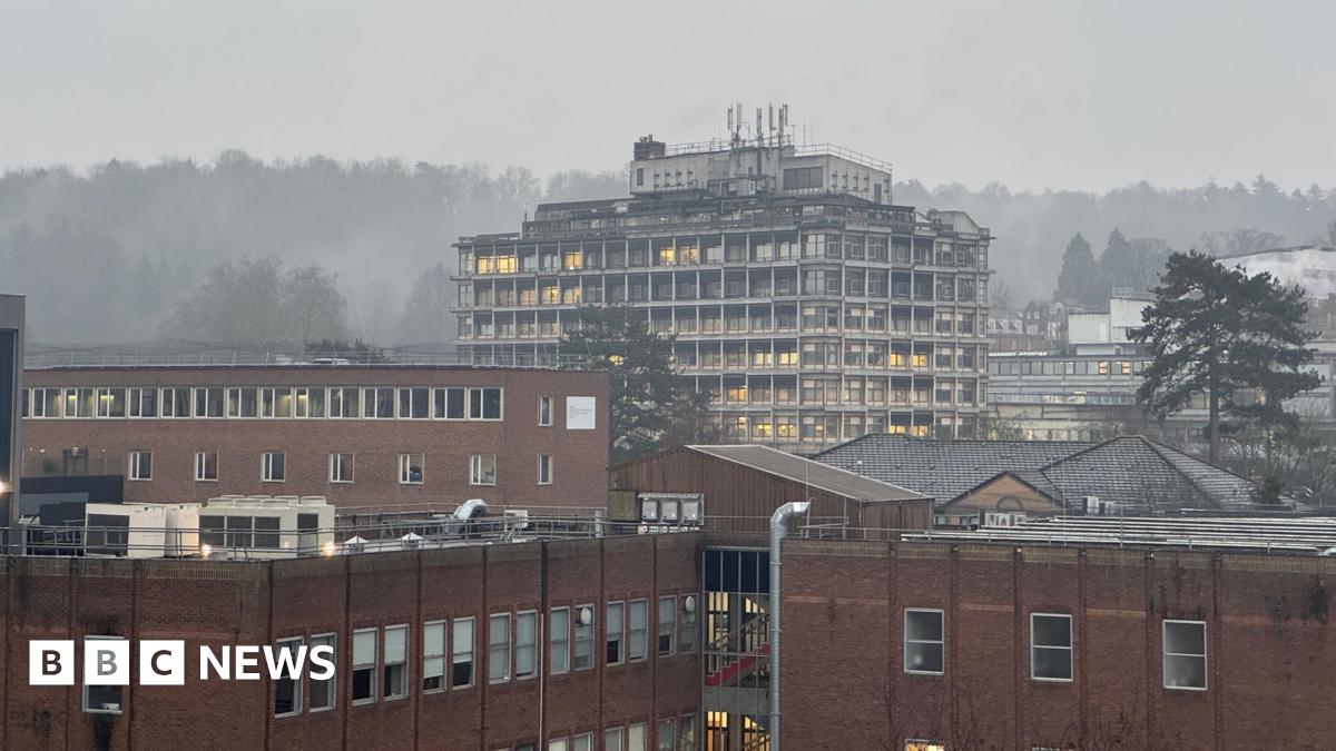 Millie Bobby Brown and David Harbour in character as Eleven and Jim Hopper. Eleven wears red shorts over grey leggings and a grey jumper. Jim Hopper wears a black leather jacket, a black beanie hat and dark trousers.They are speaking to director Frank Darabont on the set of Stranger Things. There is a vintage advertisement posted in the background with debrief on the ground.