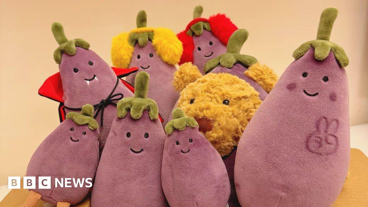 Eight Jellycat aubergine plushies of different sizes surround a teddy bear at the centre. A peace sign is drawn on the rightmost aubergine.