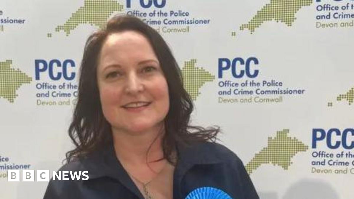 Alison Hernandez wearing a gold necklace and a navy blue top with the top of a Conservative rosette visible and standing in front of a board with the logo for PCC - Office of the Police and Crime Commissioner, Devon and Cornwall.