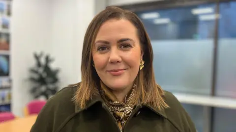 Rachel smiling. She is wearing gold flower shaped earrings, a dark green coat and khaki patterned scarf. She has shoulder length brown hair.
