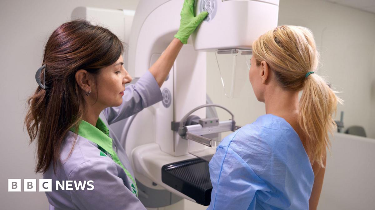 A woman stands in a hospital gown undergoing a mammogram. A medical practitioner stands beside her in a black top.