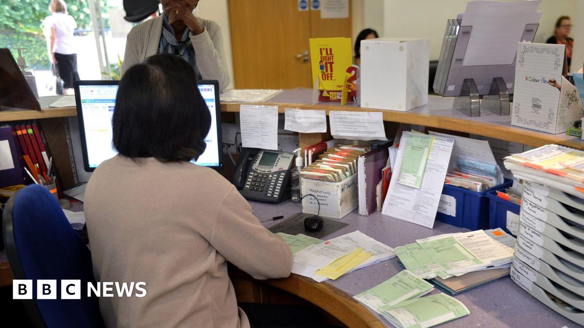 A GP surgery reception. The receptionist has her back to the camera and is talking to a female patient who is standing at the desk. The desk has a phone, PC, prescriptions and filing systems. Beyond is a waiting room where two women are sitting.