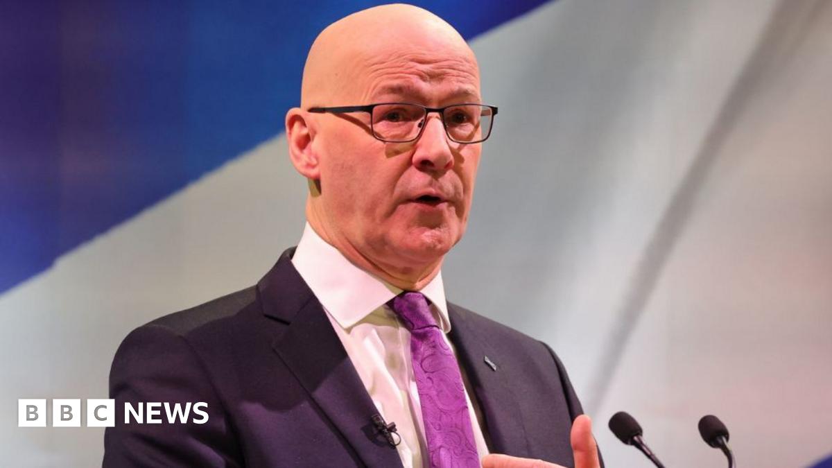 John Swinney, who is bald and wearing glasses, stands at a podium in front of a saltire. He is wearing a dark suit, white shirt and purple tie.