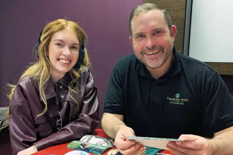 Two people sitting at a red table covered with printed wildlife photographs, including images of animals and landscapes. One person is wearing a maroon leather jacket over a patterned top, and the other is wearing a black polo shirt with a green logo and text that reads 'Touch the Wild.'