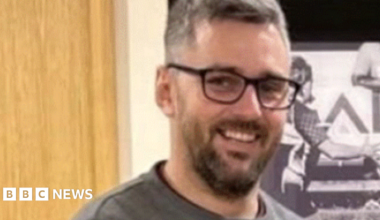 A blurry image of Craig Hurcombe who is smiling at the camera. He has a dark beard and glasses