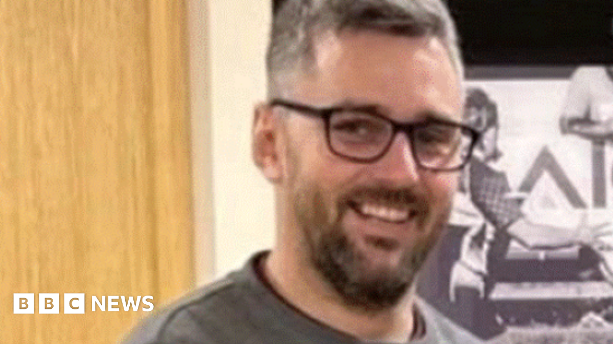 A blurry image of Craig Hurcombe who is smiling at the camera. He has a dark beard and glasses