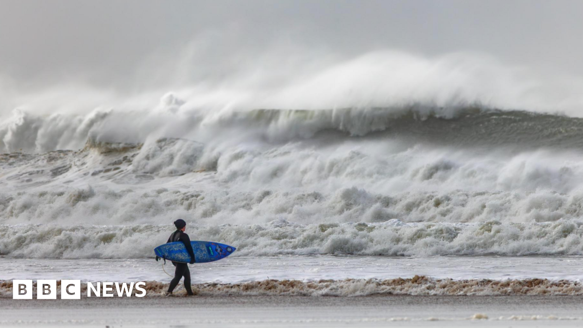 The picture shows a lone surfer standing on the shoreline, holding a blue surfboard and looking out towards huge, churning waves. The sea is rough and wild, with towering white‑topped swells crashing in the distance. The sky looks grey and stormy.