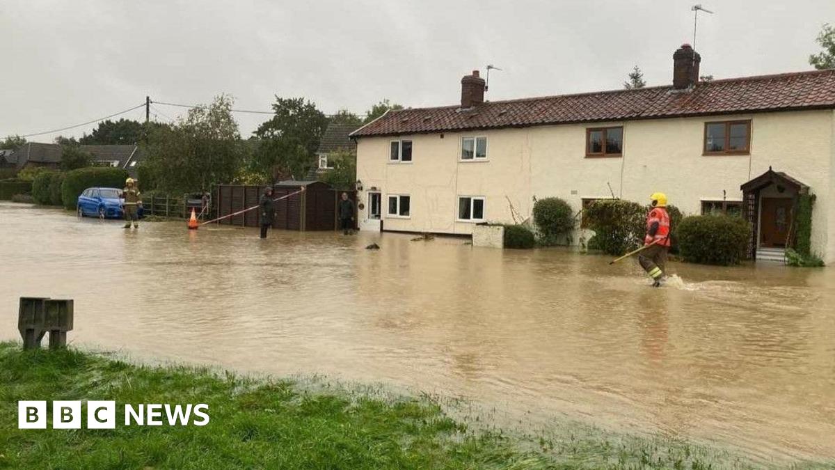 A row of cream-coloured houses looking out over a wide body of water, which goes up to the front doors. A firefighter wades through the water while another firefighter is on the left.
