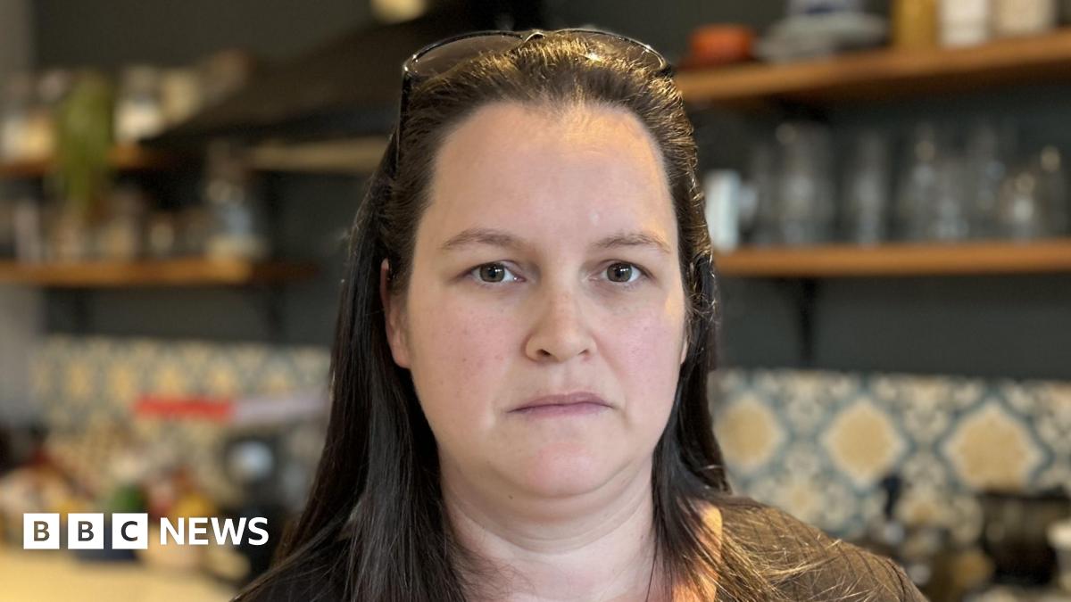 A woman stands in a kitchen. She has long brown hair and is wearing a brown top