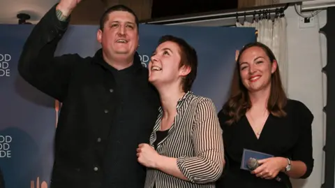 Getty Images Chef Gareth Ward stands on a small stage in front of a “The Good Food Guide” backdrop. He raises an award plaque above his head while smiling, his guest, a woman, looks up at it, smiling, and a woman on the right smiles while holding a microphone and a card.