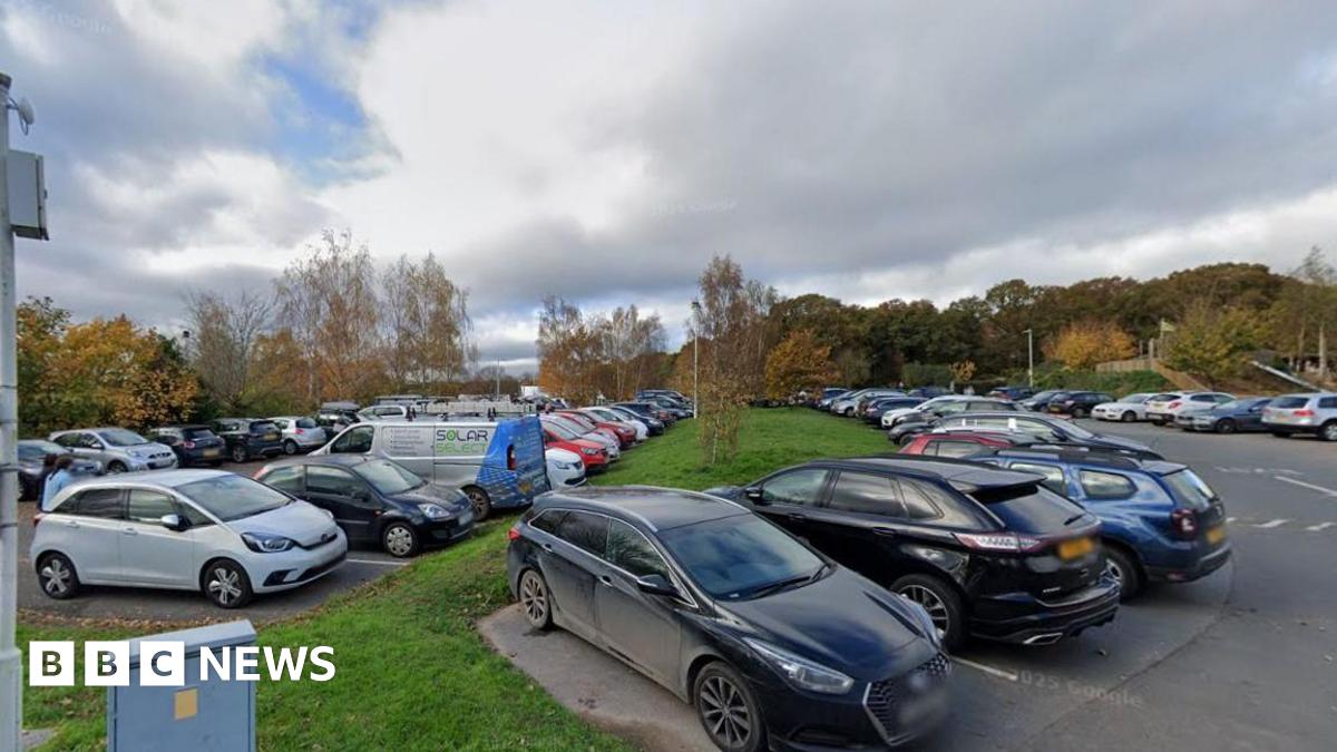 A large car park that is full of cars. There is a small strip of grassland in between two sections of the car park with trees in the distance.