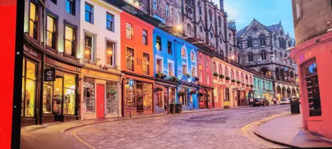 Li Hong A row of shops on a cobbled Edinburgh street. The shops and flats are various colours, such as white, blue and pink