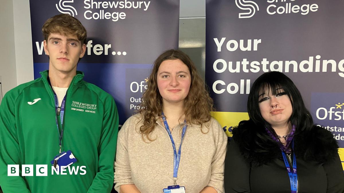 A tall youth with blonde curly hair and a green hoodie next to a young woman with long blonde wavy hair and a beige jumper and a shorter young woman with black hair and a black top