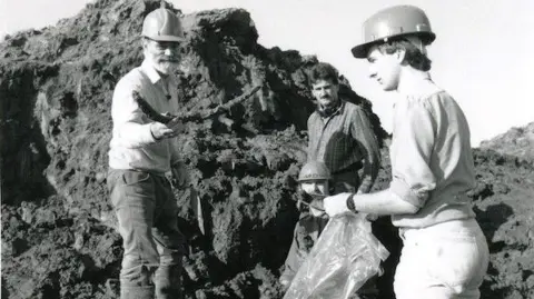 Shropshire Museums A black and white image of four men in a quarry. There is a large mound of clay and mud in the background. Three of the men have hard hats on. One man, with a light coloured beard is holding a curved bone towards another man, who holds out a plastic bag