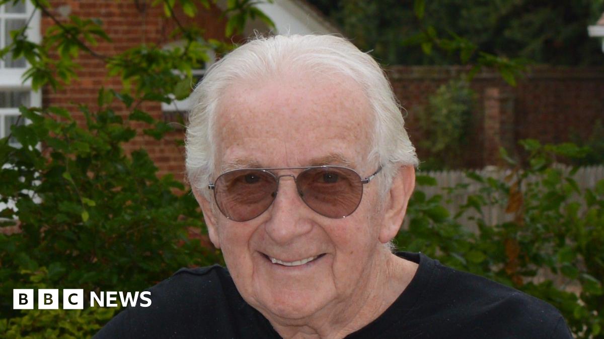 Ian Chatten smiles at the camera. He has short grey hair and wears sunglasses along with a black T-shirt. He is standing in front of a building with bushes and trees behind him.