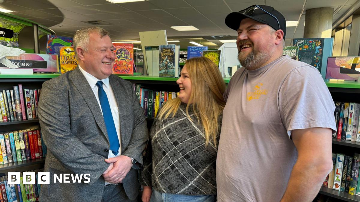 Amy and Ben Grainger stand smiling in a library, with book cases visible behind them. He's wearing a light brown tee-shirt and her a plaid top. They are standing next to Councillor Nick Lakin in a grey suit and blue tie