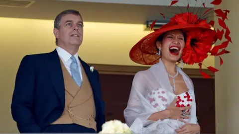 Getty Images Andrew Mountbatten-Windsor, wearing a black morning coat and a taupe waistcoat with a pale blue tie, looks in the distance with his hands in his pockets at Royal Ascot, while beside him Goga Ashkenazi, who wears a large red hat and a white dress with red spots, appears to laugh uproariously