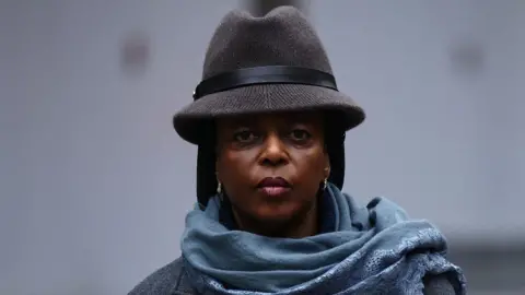 Getty Images Diezani Alison-Madueke arrives at Southwark Crown Court on 26 January. She is looking at the camera, and wearing a blue scarf and brown hat