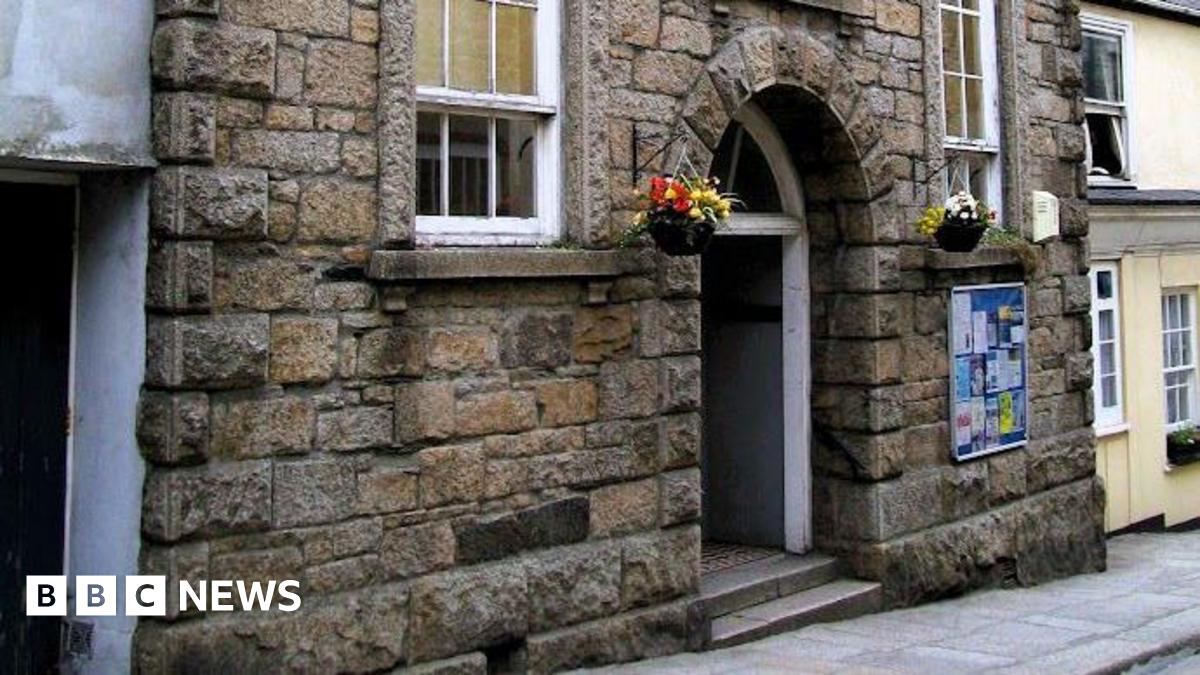 A brown brick building with a large arched door. There are large windows either side of the door. There are flowers in baskets.