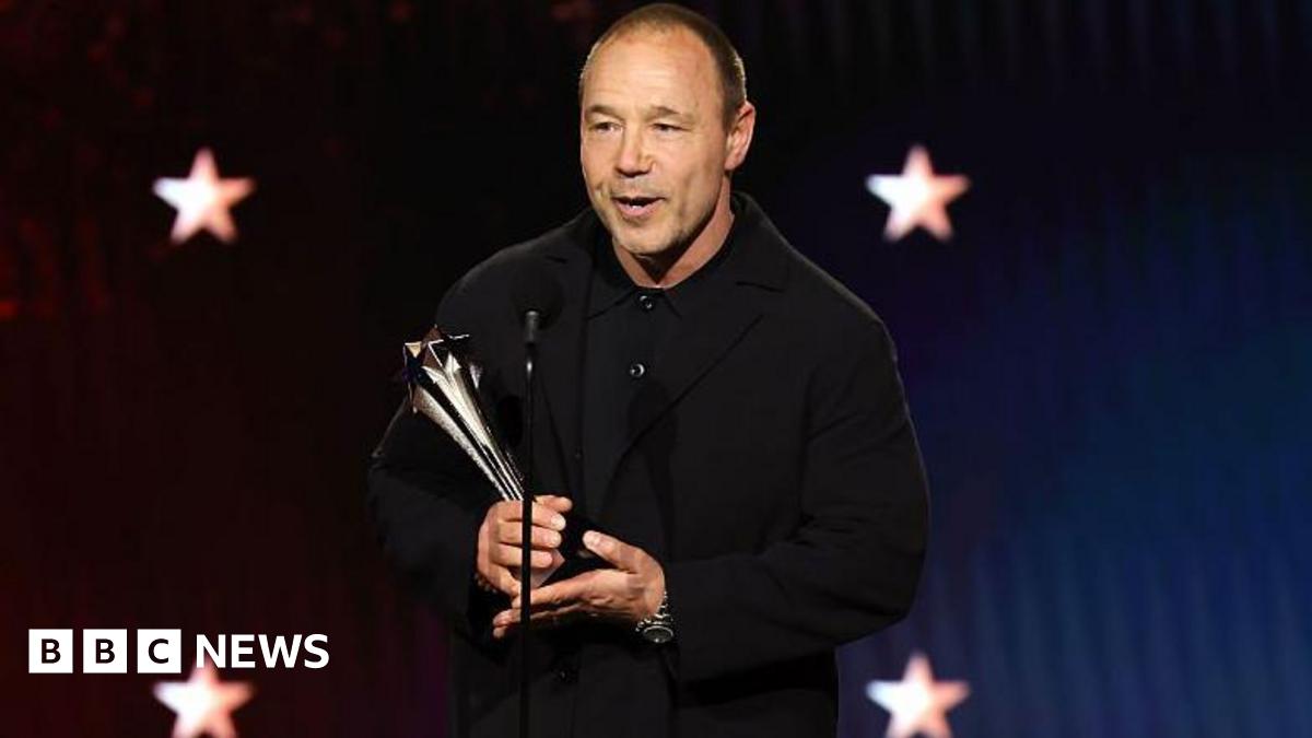 Stephen Graham in dark clothing holding a silver trophy. He is talking into a microphone in front of a dark background with four white stars on it.