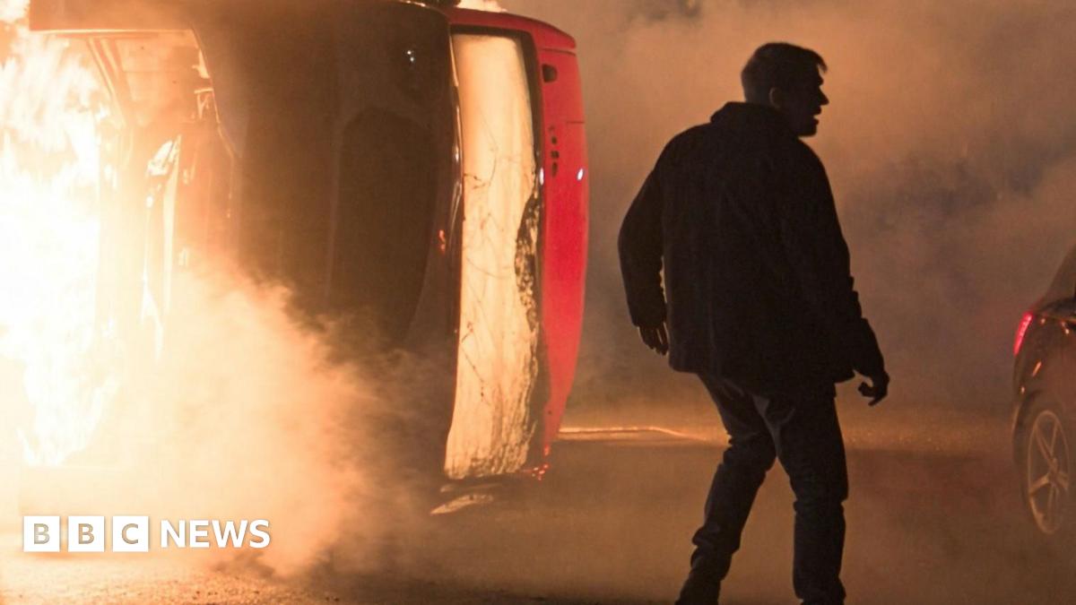 The figure of a character standing in silhouette on a road at night in front of a burning coach and a crashed car, in a scene from Corriedale