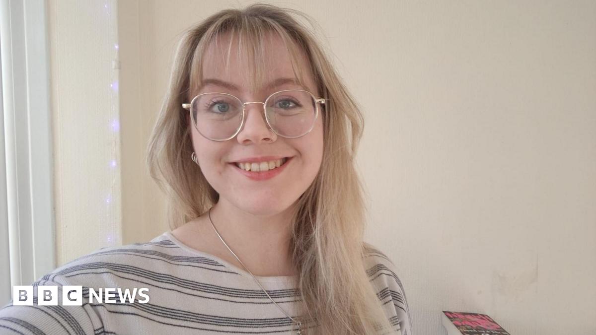 Catherine smiles at the camera, with long blonde hair, glasses, and a white top with black stripes. In the background are fairy lights and a pile of books.