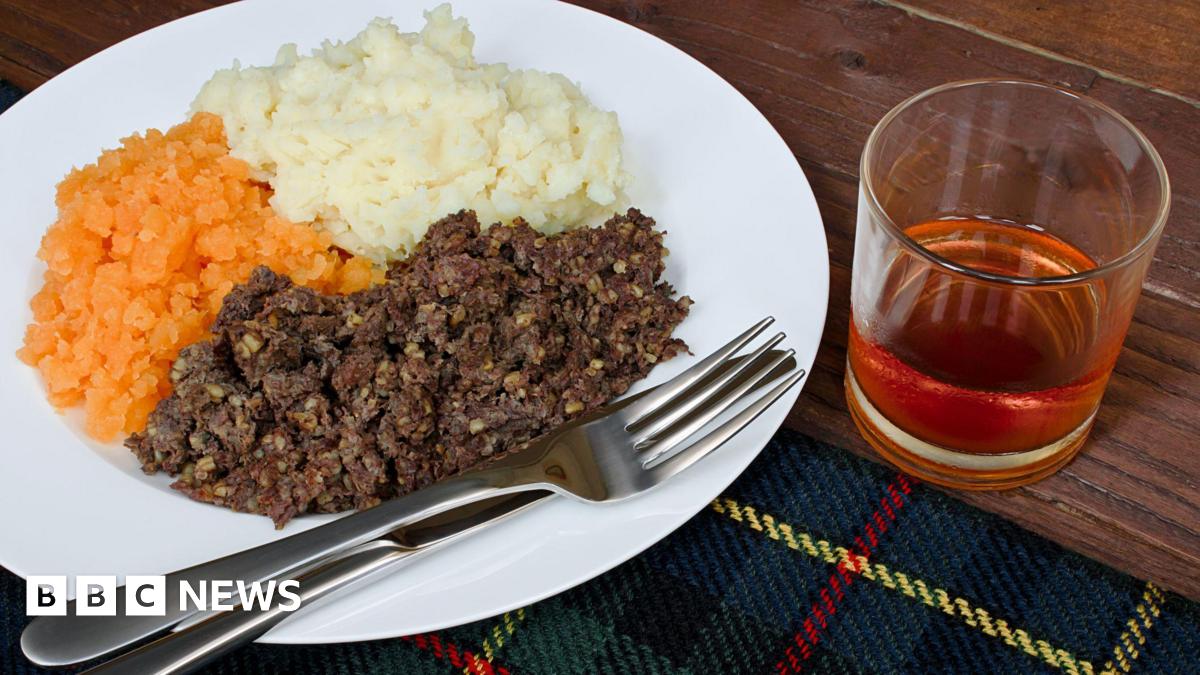 A white plate with a knife and fork, with brown haggis, mashed orange turnip and white mashed potatoes sitting on a tartan cloth on top of a table. There is a glass of whisky beside the plate.