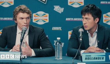 Rachel Reid, a woman with short, curly brown hair, leans on her left hand and smiles. She is wearing a dark red top and a gold necklace.
