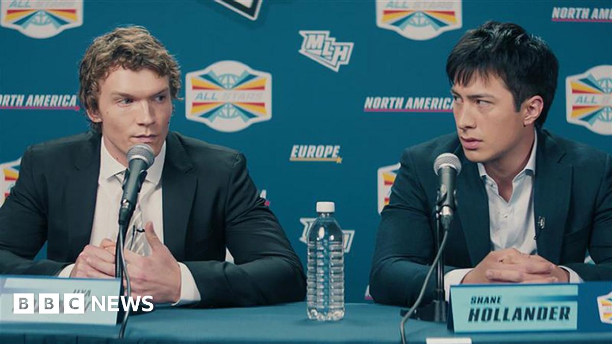 Rachel Reid, a woman with short, curly brown hair, leans on her left hand and smiles. She is wearing a dark red top and a gold necklace.