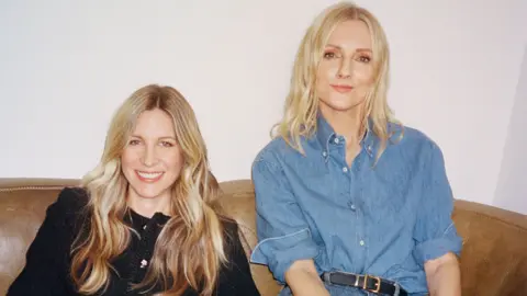 BBC Two women with blonde hair smiling, one wearing a black jacket and the other a blue button up shirt.