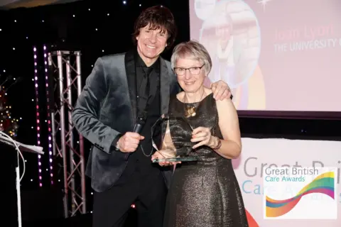 Great British Care Awards Joan Lyon, a 75-year-old woman, with grey bobbed hair and glasses, wearing a brown and gold dress. She is receiving a Great British Care Award from the entertainer Steve Walls, who is wearing a black velvet jacket and a black shirt and tie