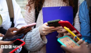 Six young people standing in a group holding smartphones of different colours.