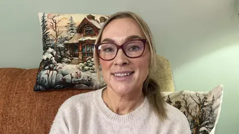 BBC A woman wearing a stone-coloured jumper. She is sitting on a couch with a light brown blanket. Behind her head is a Christmas themed cushion and her left-hand shoulder is another Christmas themed cushion 
