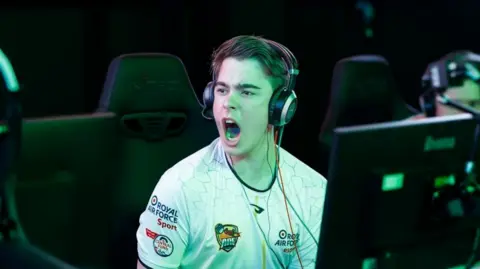 BFBS A young man wearing a gaming headset and a white esports team jersey roars in celebration as he looks over at a fellow player. He's bathed in a green glow from the nearby computer monitors. His jersey bears the badge of his team and "Royal Air Force Sport" logos.