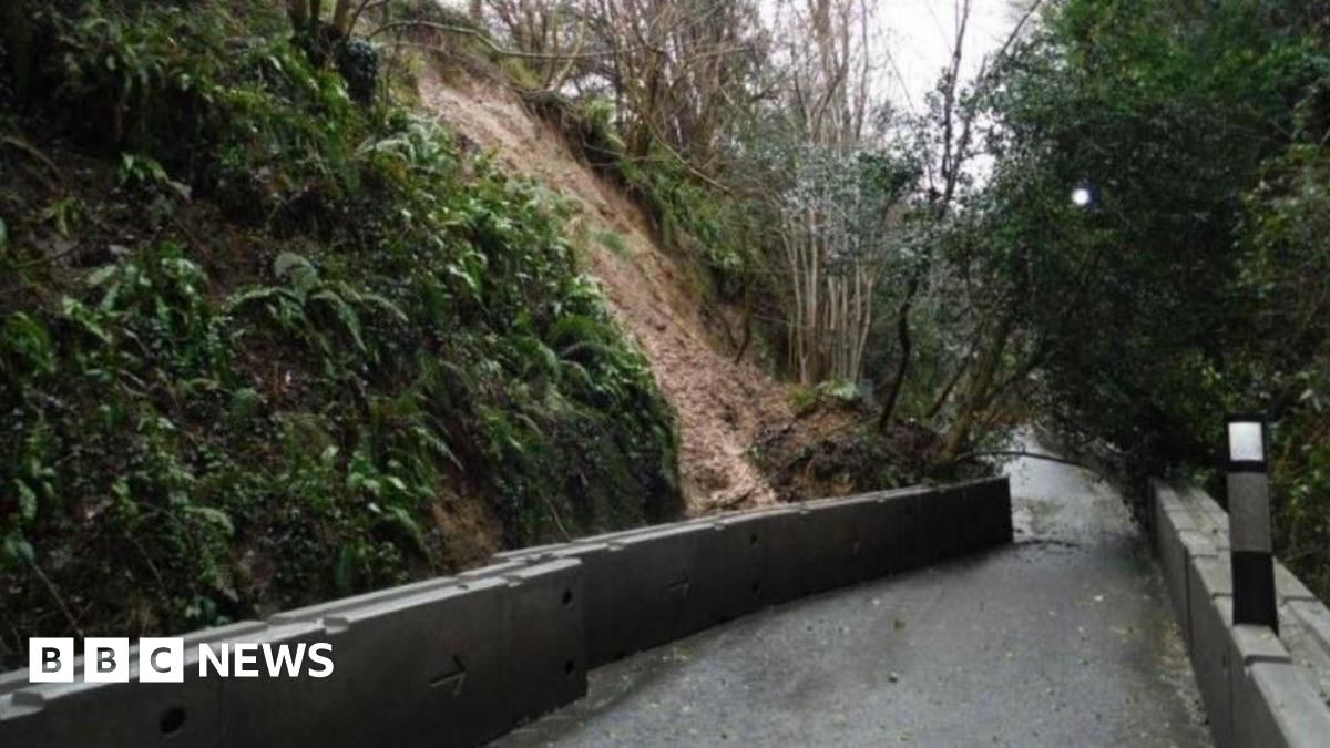 A photo of the C13 road with high, steep banks either side covered in trees and vegetation. The bank on the left side has collapsed and a long, foot-high concrete retaining barrier has been pushed into the road by the fallen soil, completely blocking it.