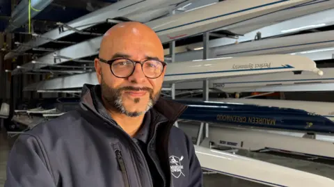 Ben Schofield/BBC Omar Terywall, to the left of centre frame and looking directly out of the image. He is smiling slightly, has a dark, neatly trimmed beard, flecked with grey on his chin, and has on circular, black-framed glasses. He is bald. He is wearing a dark blue, collared top, with a white 'Cambridge Rowing' insignia on the right. Behind Omar are several sporting rowing boats mounted on racks inside a boathouse.