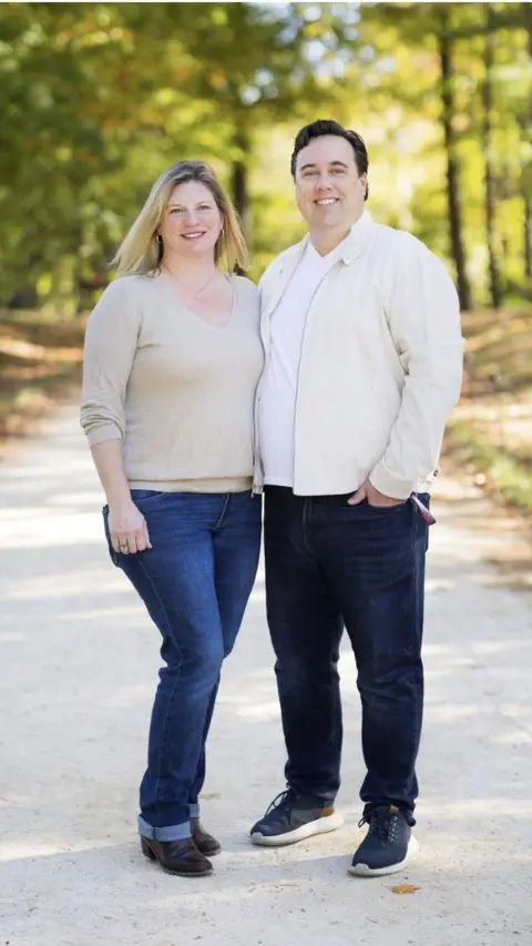 Phil Barr stands with his wife on a path in a local park, with green trees seen out of focus in the background. Phil and his wife are wearing cream coloured tops and jeans.
