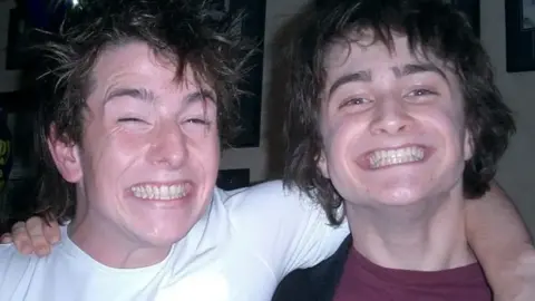 David Holmes David and Daniel when they were younger, smiling at the camera. David is in a white T-shirt and Daniel is in a red T-shirt. Both are pulling large manic grins, and they have messy dark brown hair.