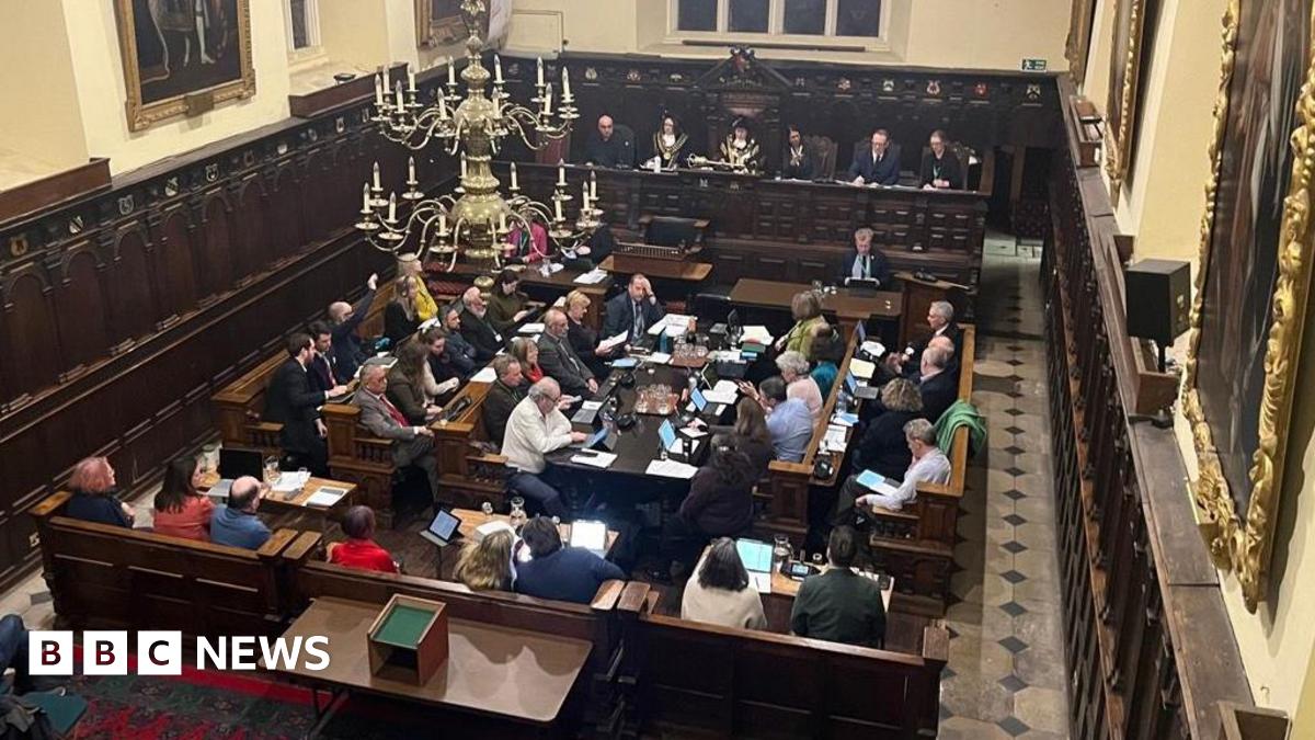 Millie Bobby Brown and David Harbour in character as Eleven and Jim Hopper. Eleven wears red shorts over grey leggings and a grey jumper. Jim Hopper wears a black leather jacket, a black beanie hat and dark trousers.They are speaking to director Frank Darabont on the set of Stranger Things. There is a vintage advertisement posted in the background with debrief on the ground.