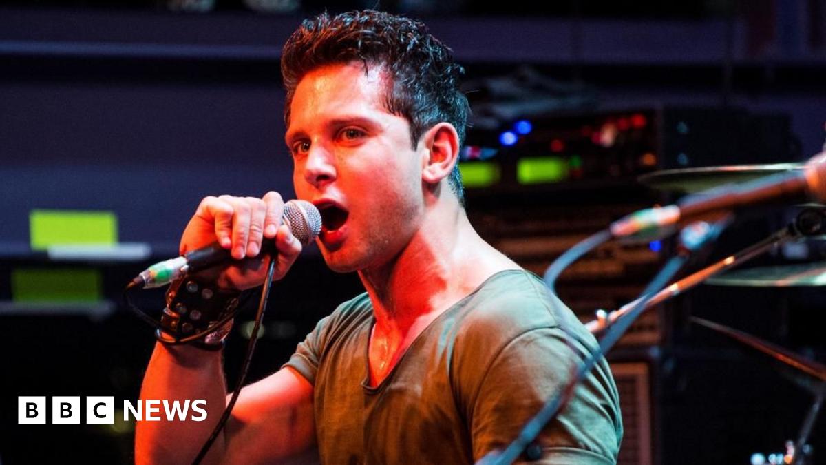 Man with brown hair sings into microphone on stage with red lighting shining down on him. He wears a tan t-shirt and black punk wristband.