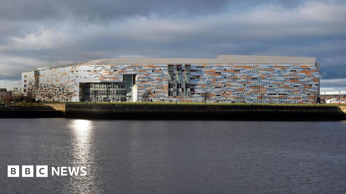 A general view of Middlesbrough College taken across the dock, with the water in the foreground. The large building is clad with grey, blue and orange material creating an irregular pattern.