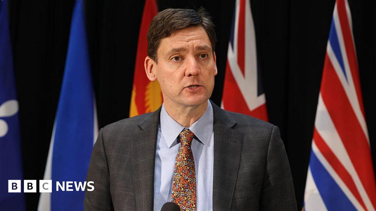 David Eby speaks into a microphone while behind him are various flags from each Canadian province,