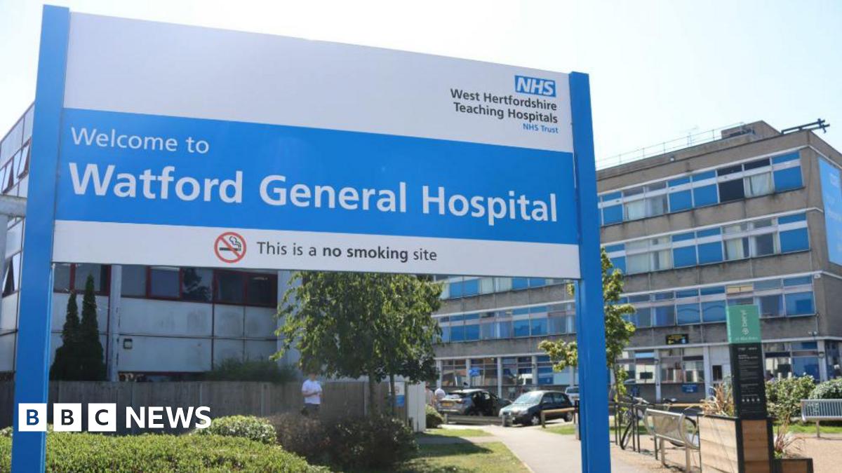 A large blue and white sign is in the foreground saying Welcome to Watford General Hospital" with the NHS logo above it. The hopsital - a concrete block of 4 storeys -is behind.